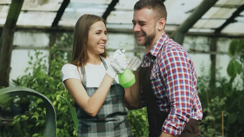 Happy young florist couple in apron have fun while working in greenhouse. Laughing woman spray water in husband face indoors